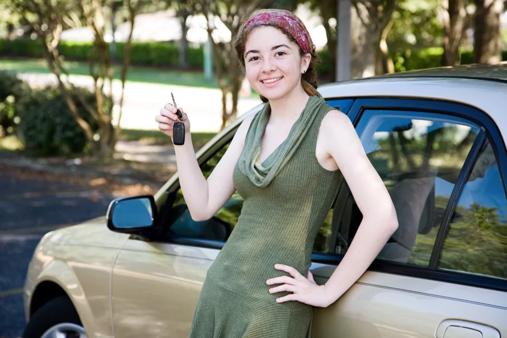 A happy young woman by her car holding up her car keys after getting her First Car Loan approved and buying a car.